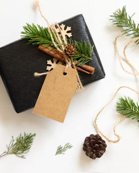 Black present with Christmas vertical gift tag, fir tree branches, pine cones and holiday decorations, top view. Winter composition with blank label Mockup, copy space, top view