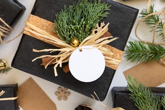 Black wrapped present with Christmas gift tag with fir tree branches, pine cones and holiday decorations, top view. Rustic winter composition with blank Gift tag Mockup, copy space, close up