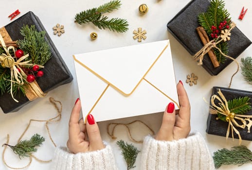 Hands with envelope between Christmas decor, black gift boxes and green fir branches top view. Blank envelope mockup, flat lay, copy space. Christmas or New Year Holiday greeting
