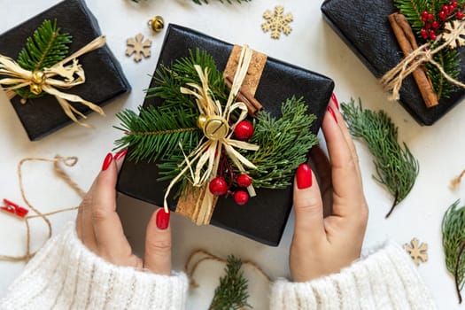 Hands with Christmas present decorated with fir branches, berries and bells, top view on white table. Winter composition, woman with a gift box
