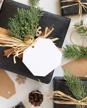 Black present with Christmas gift tag, fir tree branches, pine cones and holiday decorations, top view. Winter composition with blank label Mockup, copy space, top view