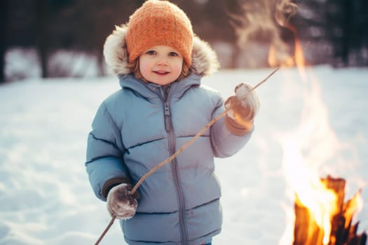 Child playing outdoors on winter day. Smiling little child is standing with stick by campfire while winter picnic. Enjoying winter holiday. Kid wearing winter clothes. Snowy winter season. Winter vacations