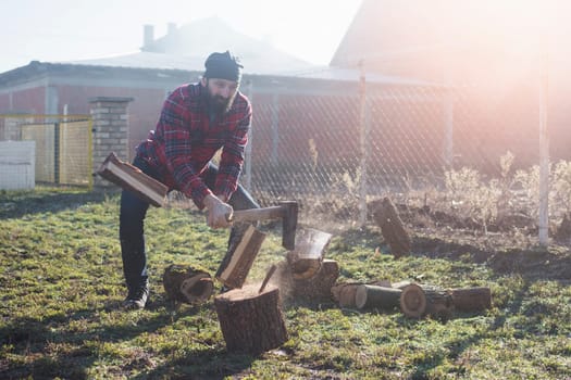 The bearded lumberjack worker or woodcutter is cutting wood or firewood with axe outdoors.
