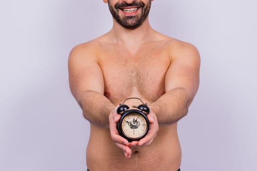 Middle-aged white man looks on an alarm clock in hand on grey background, copy space for text