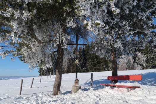 Little red bench under some snow covered trees on a sunny winter day with cross saying god bless our land, Wertach, Allgäu, Germany 2022