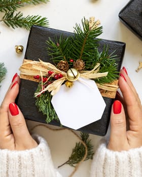 Hands with Black present with gift tag, fir tree branches, pine cones and Christmas decor, top view on white table. Winter composition with blank label Mockup, copy space