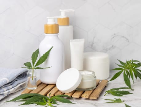 Opened cream jar with blank lid near green cannabis leaves on wooden tray in bathroom near other bottles, close up, mockup. Organic skincare beauty products, natural CBD cosmetics