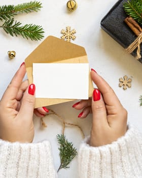 Hands with card and envelope near gift boxes, fir branches and Christmas decor, top view on white table. Winter composition with blank card, Mockup, copy space