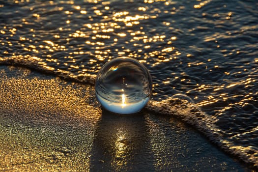 A glass ball lies in the waves on the sandy beach, the sea and the setting sun are reflected in the ball