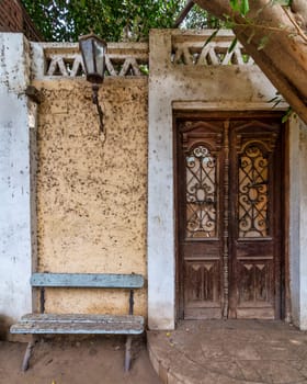 Weathered wooden blue bench sits invitingly beside of a classic wooden decorated grunge door. A vintage metal lamp hangs above the bench, adding to the nostalgic charm of the scene