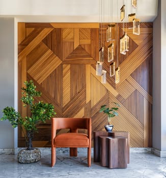 Simple and elegant shot of a modern style orange chair and coffe table in front of a decorated wood cladding wall, with hanging lights and a potted plants