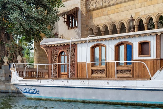 Vibrant weathered wooden antique boat, rests peacefully on River Nile infront of a Mamluk style Palace of Prince Naguib Hassan Abdallah, Dahab Island, Cairo, Egypt