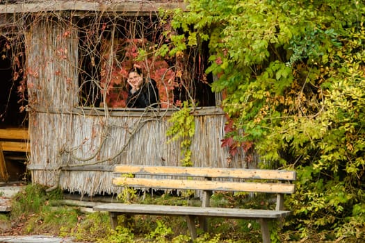 A young beautiful girl looks out of the window of an old abandoned house in the forest overgrown with plants.