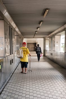 Malaysia - July 27 2023: Elderly in Yellow With a Walking Cane Crossing Sheltered Bridge