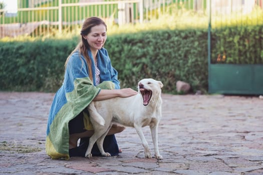 Candid portrait of young woman kneeling outside on stone ground, covered with blanket, caressing stray dog yawning next to her