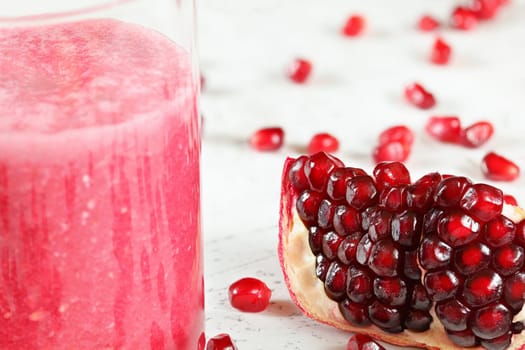 Portion of pomegranate fruit, gem like berries scattered on white board, glass with pink smoothie next to it