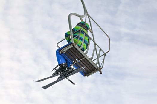 Looking up the skier in green and blue jacket, sitting on simple chairlift, sky with small clouds in background