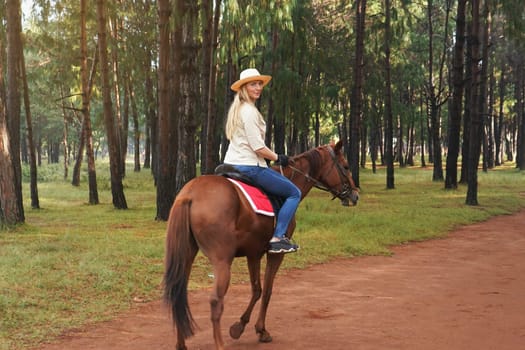 Young woman in shirt and straw hat, riding brown horse in the park looking back over her shoulder, blurred background with trees
