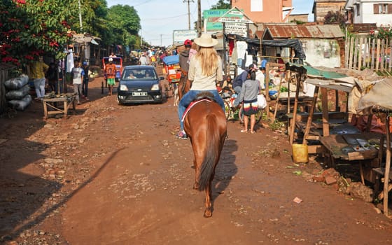 Antsirabe, Madagascar - April 26, 2019: Young woman in straw hat rides on her horse through morning market in Antsirabe, stalls on both sides, car coming in opposite direction, view from her back