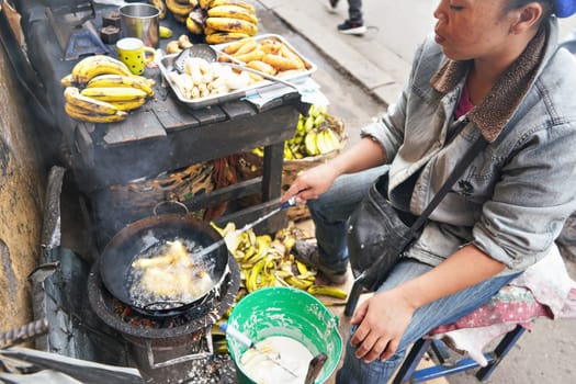 Antananarivo, Madagascar - April 24, 2019: Local Malagasy woman frying bananas in oil at her stall on market next to main road. Fresh food prepared on street is common sight in Madagascar