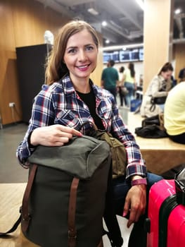 Young woman in shirt waiting for her transfer, with her backpack and luggage near her, looking into camera, smiling, blurred station in background