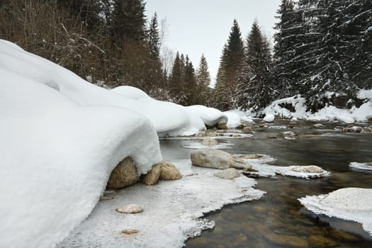 Morning on frozen river in winter, low angle photo, focus on ice patterns and stones, with coniferous trees and rising sun in distance