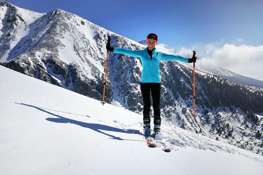 Young woman in light blue jacket standing, smiling happy with mountaineering ski and poles, hands spread, sun shines on snow, bright sky over mountains behind