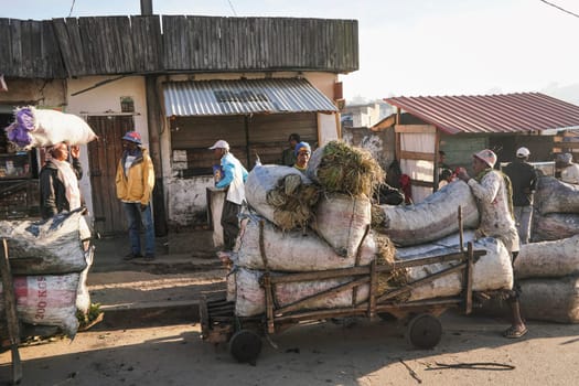 Fianarantsoa, Madagascar - May 06, 2019: Unknown Malagasy man pushing cart with bags full of straw and grass, to be sold at local market on sunny morning. More people near their stalls in background