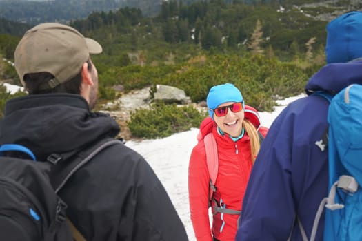 Three hikers wearing jackets, mountains and forest partially covered with snow, young woman in red jacket, blue cap and sunglasses looking to other two, smiling happily, blurred trees at background