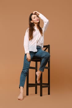 A young adult woman with long, brown hair sits casually in a studio against beige background. She is wearing jeans and poses for the photo shoot surrounded by furniture.