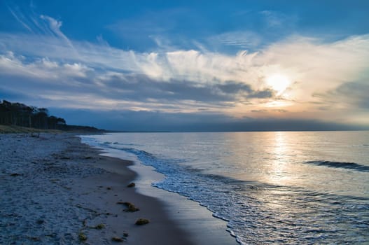 Sunset on the west beach on the Baltic Sea. Waves, beach, cloudy sky and the last rays of sunshine on the coast. Landscape photo