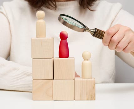 Woman holding a magnifying glass and wooden men on a white table. Personnel recruitment concept, talented employees. Career advancement