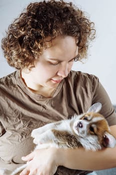 Tenderness and care. Young woman in dark beige cotton T-shirt lovingly hugs cute little Welsh corgi puppy. Dog food. Pets. Dog in family. Features of keeping puppies. Veterinary control.