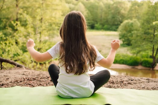 Rear view of little girl with brown long hair sitting on green sporty rug and practicing yoga in front of river. Active female sportsman in white t-shirt and black leggings meditating in nature.