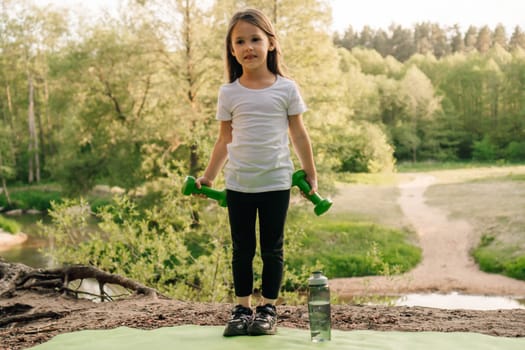 Adorable little girl holding small dumbbells in hands while standing on green mat outdoor. Junior athlete wearing white t-shirt and black leggings enjoy training on warm day, bottle lying near lady.