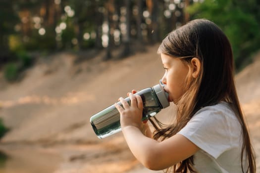 Close up of little girl with dark long hair drinking water from bottle sitting outdoor. Active kid wearing white t-shirt spending time resting on sandy beach in nature.