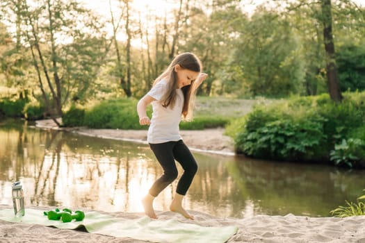 Delightful little athlete with dark long hair happily jumping on green mat on sandy beach near river. Smiling girl in white t-shirt and black leggings doing sports outdoors.