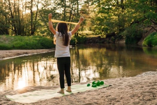 Rear view of junior sporty girl raising arms with mini band on wrists standing on green mat on sandy ground close to lake in forest. Little sportsman with brown long hair doing workout outdoor.