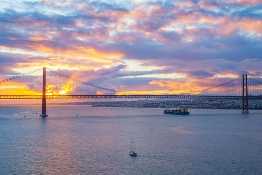 View of 25 de Abril Bridge famous tourist landmark of Lisbon over Tagus river with tourist yacht boats and cargo container ship on sunset. Lisbon, Portugal