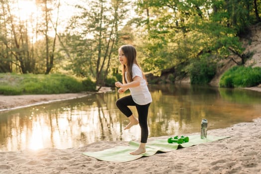 Adorable little female athlete in white t-shirt and black pants standing on green mat near river and doing sports. Energetic girl with brown long hair enjoying activity on sandy beach close to water.