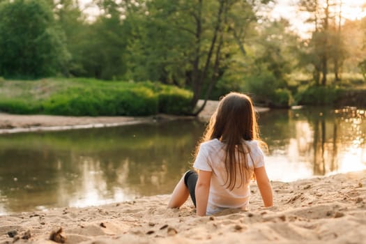 Adorable little girl with dark and long hair comfortable sitting on sand near forest. Female child wearing white t-shirt and black shorts playing with grit and having fun in nature.