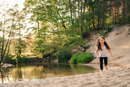 Happy, energetic girl in white t-shirt and black leggings running on sandy beach close to river. Little athlete with dark long hair doing sports and having fun in forest near water.