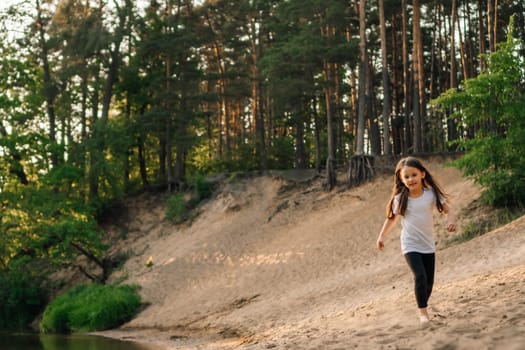Happy, energetic girl in white t-shirt and black leggings running on sandy beach close to river. Little athlete with dark long hair doing sports and having fun in forest near water.