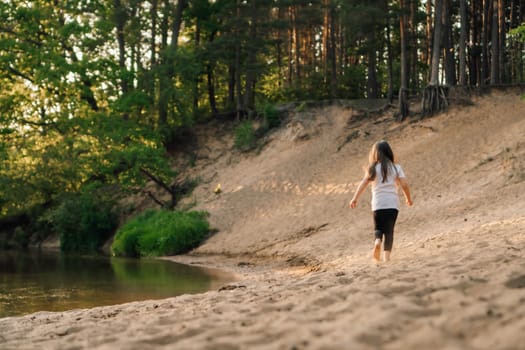 Rear view of little girl with dark, long and flowing hair running on sandy beach near river in forest. Female child in white t-shirt and black leggings actively spending time in nature.
