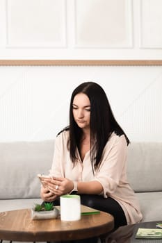 A brunette girl uses a mobile phone app, plays games, makes purchases online, orders delivery, relaxes on the couch.