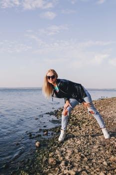 A blonde girl in sunglasses poses in ripped jeans in the afternoon on the shore of the blue sea