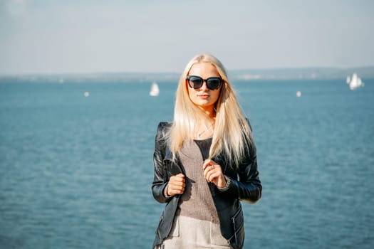 A blonde girl with long hair and glasses poses on the shore of the blue sea in the afternoon