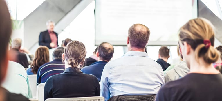 Man giving presentation in lecture hall. Male speeker having talk at public event. Participants listening to lecture. Rear view, focus on people in audience.