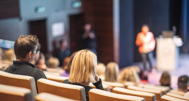 Business and entrepreneurship symposium. Female speaker giving a talk at business meeting. Audience in conference hall. Rear view of unrecognized participant in audience.