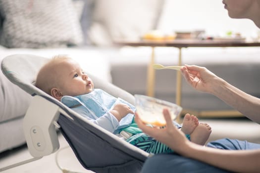 Mother spoon feeding her baby boy child in baby chair with fruit puree. Baby solid food introduction concept
